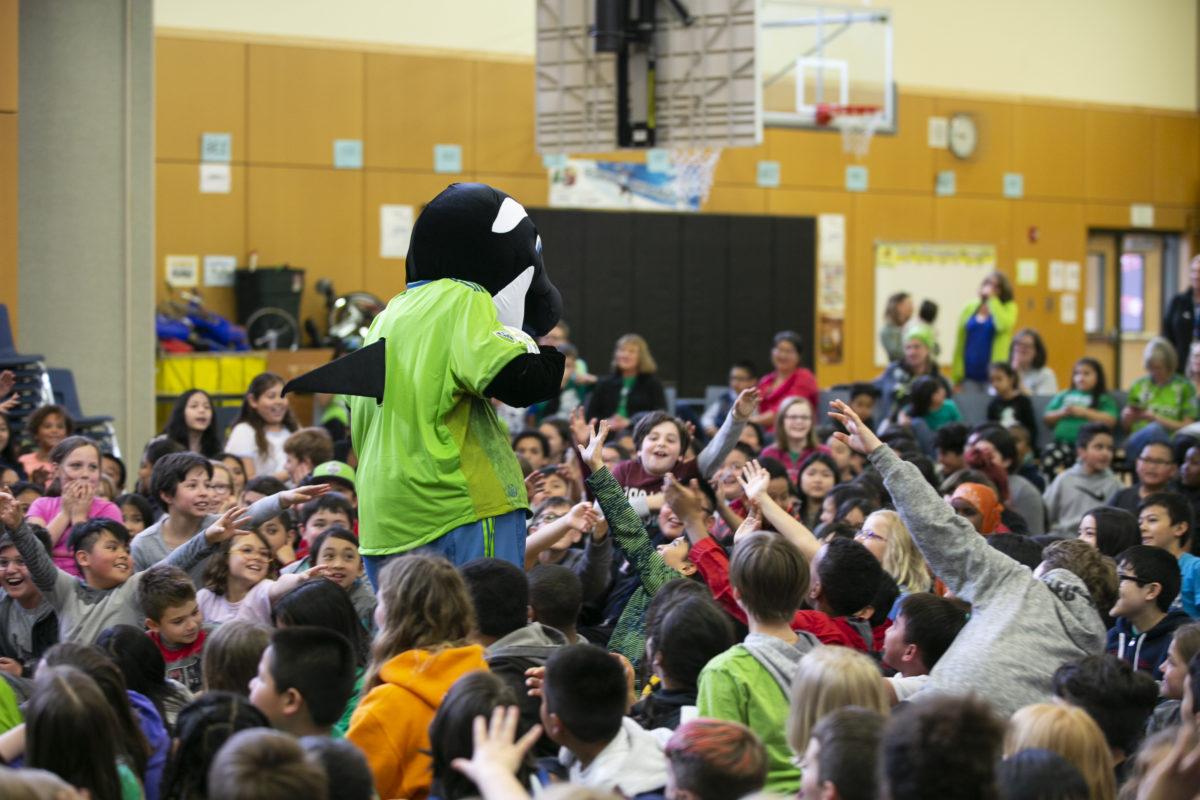 Sammy the Sounder stands surrounded by a large group of sitting children at a school assembly