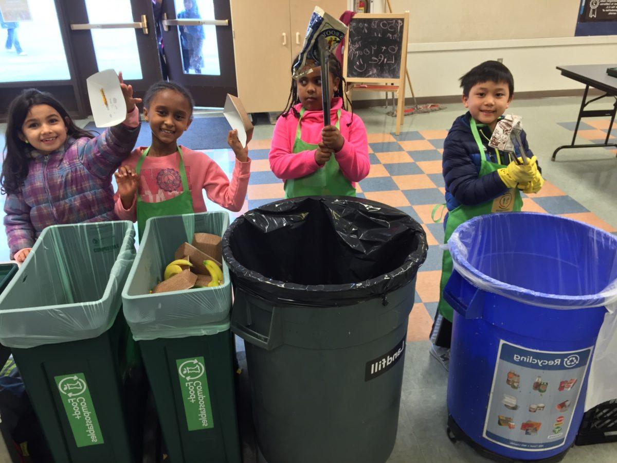 A group of 4 elementary school students poses next to waste sorting bins in their cafeteria