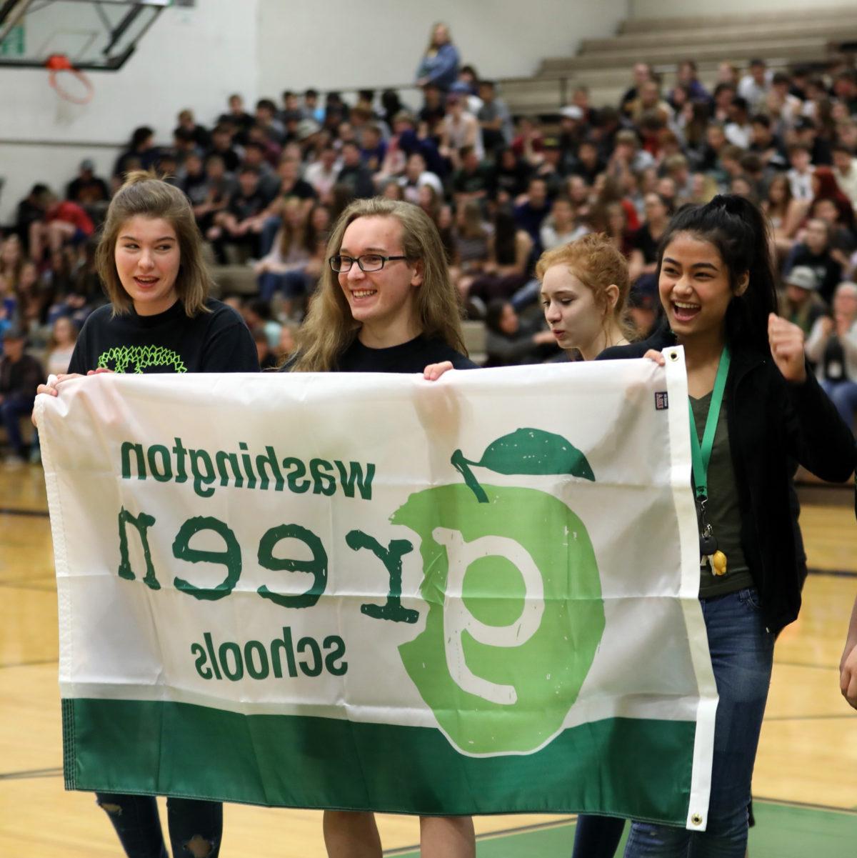 4 high school students stand excitedly behind a Washington Green Schools flag at a whole school assembly as they are recognized for their certification efforts.