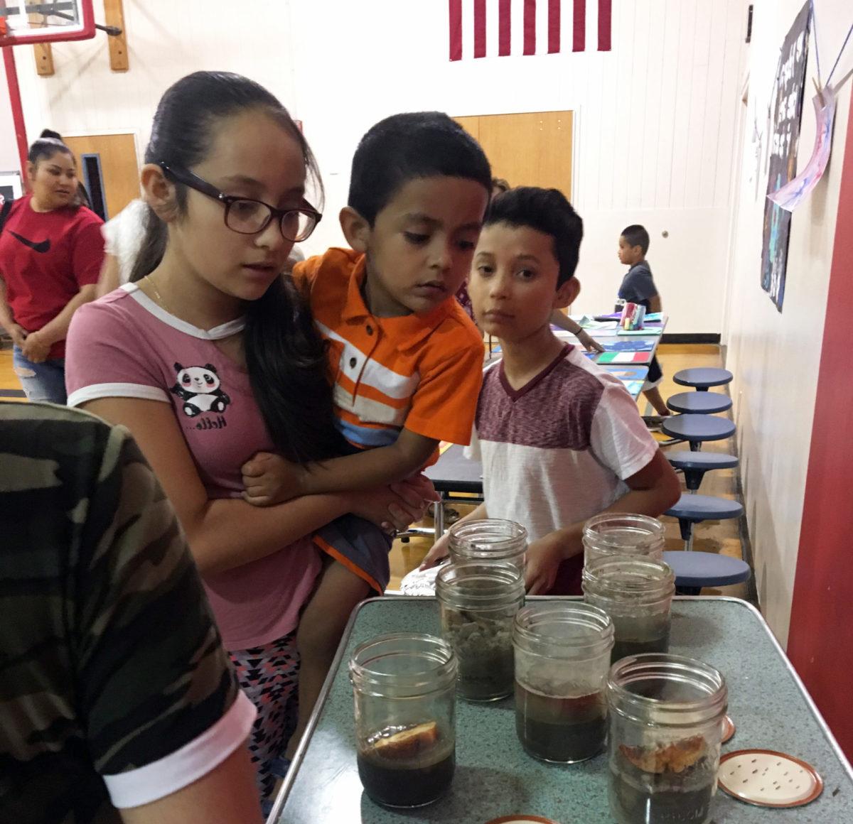 A migrant student holds her younger brother while examining mason jars filled with soil and apple slices to see which one decomposed the quickest as part of EarthGen's Zombie Guacamole program